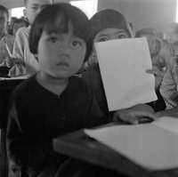 Original title: "VN Life",two girls sit in a classroom at a desk, looking at the camera. One holds a notebook over her face. There are more students in the background.