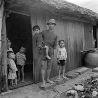 "VN Life", A man stands with five small children in the doorway of a traditional wood home with a tatched roof. There appears to be a pile of coconut husks in front of the house.