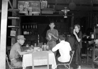 A group of Vietnamese men sit in a restaurant with a man and a woman standing in the background.