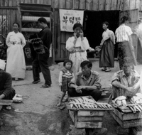 "Korea-Negs 181-3(1)-182-3(1) 5.Seoul Mkt [Market]." Two men sit on the ground selling fish from wooden crates. Behind them are people walking on a dirt path, including 3 women, a small child, and two men carrying mats. A Wooden building is in the background. From scrapbook page 23.