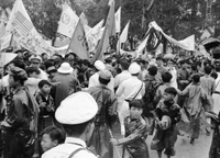 A crowd of Vietnamese people holding flags and banners. They have both American and South Vietnamese flags. Many people are wearing raincoats.