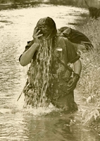 An American soldier is standing in some body of water, and empties water from his helmet. The water covers the soldiers face.