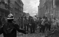 "Embassy Bombing" A crowd of people in a debri-ridden street. Most are taking pictures of the wreckage. Some attempt to help or to avoid stepping on others on the ground. Two men carry a body on a stretcher away from the scene. A fireman stands with his arms out in front of the photographer.