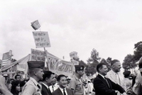 Robert McNamara and Henry Cabot Lodge Jr walk in front of a crowd holding banners and flags.