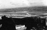 An aerial photograph of an air base and runway. Some kind of machine or gun is blocking part of the view.