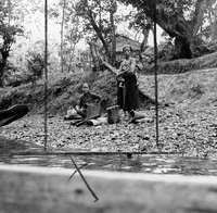 Three women in traditional clothing stand by a river. Two of the women have children teid to them with slings. Woven mats and baskets sit at their feet. The photo is marked with black lines to show how it would be cropped for publication in a newspaper.