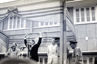 Robert McNamara waves at the crowd and Nguyá»…n KhÃ¡nh stands to the side. They are standing on a stage with microphones in front of them. Henry Cabot Lodge, Jr., appears to be standing off to the side.