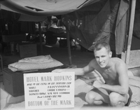 An American man poses next to sign outside a tent.  The sign reads "Hotel Mark Hopkins, Home of the Cuvs[?], of the Fighting 8th. Norvi, J.B. Pennington III, Buscher, Corrie C. Cohins, D Lowell Growe, Wm Geo Sirahur, and A Rankin Ruiz PhD, the Bottom of the Mark."