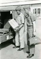 Two American soldiers are loading supplies into their Jeep. One is holding a box and the other named Hibbs holds a medical bag.