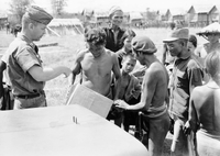 An American soldier named Hibbs is handing out supplies to villagers. Two villagers hold a cardboard box.