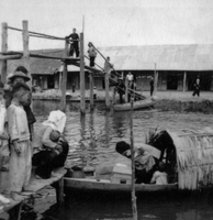 A Vietnamese boat approaches a dock or landing. A woman holding a child holds the boat steady, as the remaining people on the boat prepare to disembark. Children are looking on from the dock.