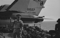 "7th Fleet" An American man wearing a headset sits beside what may be artillery on the USS Kitty Hawk. In the foreground stands an officer giving the thumbs-up sign.