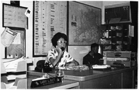 Beverly Keever  sits at a desk talking on the phone in the Saigon AP Office. The wall behind keever is covered in calendars, maps, and schedules. The desk next to Keever has a small sign reading "Larry L. Rutt LÃºc Giáº­n Thi Dá»§ng NÃ³i[?]." In the background, a man sits near a pidgeonhole cabinet.