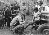 An American soldier named Hibbs is bandaging a woman's leg as many other villagers are watching. The woman sits on the bumper of a Jeep.