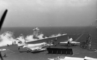 Men working on an aircraft carrier in open water. Three Naval planes are still on the runway. Two have just taken off.