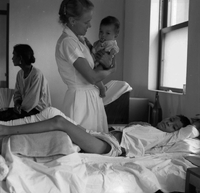 A Vietnamese child lays in a bed with their leg propped up as an American nurse holds a baby in the background.