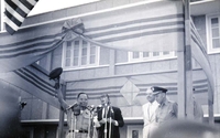 Nguyá»…n KhÃ¡nh holds up his hat as Robert McNamara looks at his speech cards in the background. They are standing on a stage with microphones in front of them. Henry Cabot Lodge, Jr., appears to be standing off to the side.