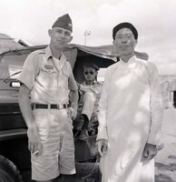 An American soldier is posing next to a villager for a photo. There is a Jeep in the background.