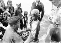 An American soldier named Hibbs examining an elderly man's hand or arm. Villagers watch in the background.