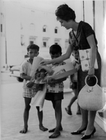 Beverly Keever with two newsboys on a sidewalk in Saigon. Keever takes a newspaper from one of the boys. She is carrying a hanbag and a wrapped gift. She is dressed in a fashionable dress. A third boy walks through the background of the photo, staring at the camera. This photo was taken by a reporter who was writing about Keever for an article in Mademoiselle. It is unclear whether the article was ever published