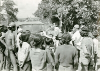Two American soldiers are examining villagers near their parked Jeep. Many villagers are gathered around to watch