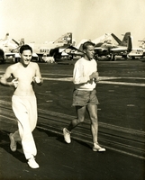 "An empty flight deck on the Aircraft Carrier America provides ample room for exercise. Ensign Nedum Haley, Atlanta, Georgia and Corporal Alan Kirkman, Lawrence, Massachusetts, both of the carrier division two staff, run laps to keep in shape while the Norfolk-based carrier is employed to the Tonkin Gulf." Two American men jogging on what appears to be an aircraft carrier.  There is a row of jets parked in the background.