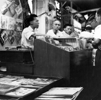 "Sarawak." A stall in a Sarawak night market selling LP records and some printed materials (possibly small books). There are 3 vendors and 2 customer in the photo. The stall is decorated with LP covers and there is a record player behind the case. From scrapbook page 13.