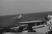 "7th Fleet" A row of Navy airplanes sitting on an aircraft carrier in open water. One airplane has just taken off.