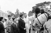 Robert McNamara and Henry Cabot Lodge Jr walk in front of a crowd holding banners and flags. There is press in front of him taking photos.