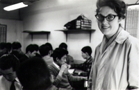 Beverly Keever stands in a classroom in the foreground smiling at the camera. In the background are a group of young Japanese men working at tables.