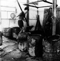 "Indonesian Arts and Crafts Negs [Negatives]." Two Indonesian men stand in a room with barrels of dyeing fabric. One man looks at the camera and the other lifts the fabric in a large sieve. From a scrapbook page 10.