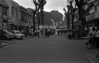"Unidentified", Photo of a protest. A large crowd of Vietnamese people walk in a tree lined street with banners. The street is lined with small shops, and pedestrians have paused to watch.