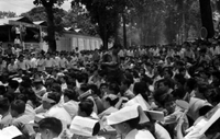 "Unidentified", a group of protesters, largely young men, sit in a city street. They appear to be listeneing or watching something off camera. Some shield themselves from the sun with pieces of paper.
