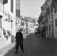 "Macau" A woman walks on a cobbled street towards the foreground, while a group of children play in the background.