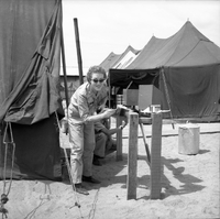 Two American women paint a fence post as one looks at the camera.  There are field hospital tents in the background.