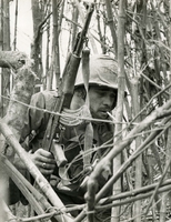 American soldier is kneeling in a field of branches with his gun. The soldier is smoking.