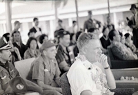 A seated Henry Cabot Lodge Jr is wearing a lei. He is surrounded by many other military members and officers, from both the Vietnamese and American militaries.