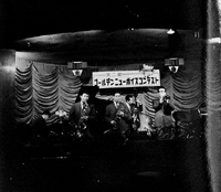 A group of Japanese men stand on stage playing instruments. There is a piano, two saxaphones, a trumpet, a bass, and a drum set. The stage is brightly lit, but the rest of the photo is very dark. There is a sign in japanese hung behind them.