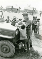 An American soldier named Hibbs is digging through his medical bag on the hood of a Jeep as a young child closely watches.
