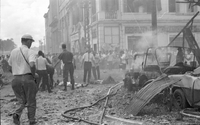 "Embassy Bombing" Two cars have collided in the steaming rubble. A crowd of people stand nearby. Men in uniform push the crowd away from two bodies lying in the rubble.