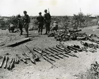 "Captured Munitions - During heavy attack on Long Binh post Jan. 31, the 9th Infantry Division's 2d Mechanized Battalion, 47th Infantry, were credited with over 200 enemy kills and 32 detainees. Weapons captured, some of which are shown above, were 26 AK-47 automatic rifles, 16 light machineguns, 24 RPG-2 rocket launchers, 12 RPG-7 rocket launchers, two .51 caliber machineguns, three 9mm ChiCom pistols, one RPK-2 light machinegun, four carbines and two 60mm mortars. Also captured were food, medical supplies, packs and clothing." Weapons and military supplies are spread across the ground, including guns, missile or grenade launchers, grenades, and rolls of cloth (possibly tents). Six American soldiers stand or kneel in the background.