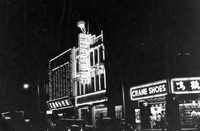 "Sarawak." View of a Sarawak street at night. There are cars on the road and buildings lit with flourescent signs and bright shop windows. One business is labelled "Crane Shoes." The other signs are in Chinese characters. One building appears to be a jewellry store, since it has a ring on its sign. From scrapbook page 13.