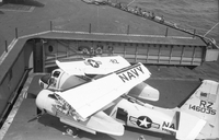 A Naval plane with detached wings on an aircraft carrier in open water. A line of American men stand along the edge of the ship.