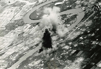 "Smoke and Dust rise from the Boi Hoa highway ferry, located 37 statute miles southeast of Vinh, during an attack with 500-pound bombs by pilots from the attack carrier USS Hancock, Aug. 27." An Aerial photograph of a bomb going off near a river in a rural area. There are many craters scattered in the landscape.