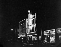 "Sarawak." View of a Sarawak street at night. There are cars on the road and buildings lit with flourescent signs and bright shop windows. One business is labelled "Crane Shoes." The other signs are in Chinese characters. One building appears to be a jewellry store, since it has a ring on its sign. From scrapbook page 13.