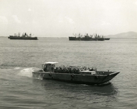"Heading Ashore - LCM-8 #870 journey's through anchorage area of Da Nang harbor enroute to Commercial Pier within the city of Da Nang. The cargo aboard the LCM-8 is combat tents slated for delivery to the Marines." An American Naval barge loaded with crates and supplies sails toward shore. There are two large Naval ships in the background.