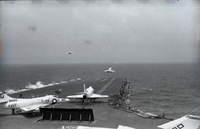 Men working on an aircraft carrier in open water. Two Naval planes are still on the runway. Two have just taken off.
