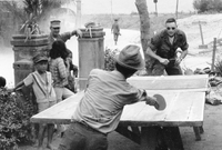 An American soldier and a Vietnamese man play a game of ping pong as others watch from the side, including some children.