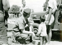 An American soldier named Hibbs kneels in front of a young Vietnamese boy near a Jeep.