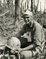 An American soldier is sitting in some brush, holding his helmet. He appears to be injured.